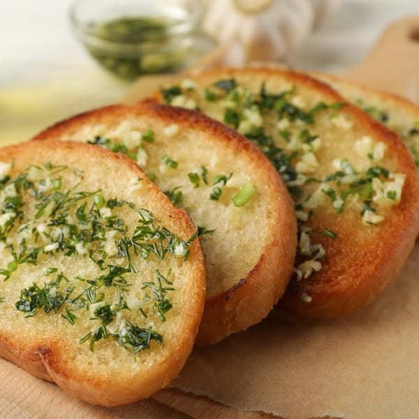 Golden-brown garlic bread slices topped with green herbs and minced garlic on a wooden board.