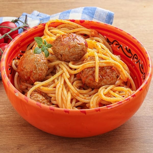 Spaghetti with meatballs and fresh basil in an orange bowl, with cherry tomatoes and a blue napkin.